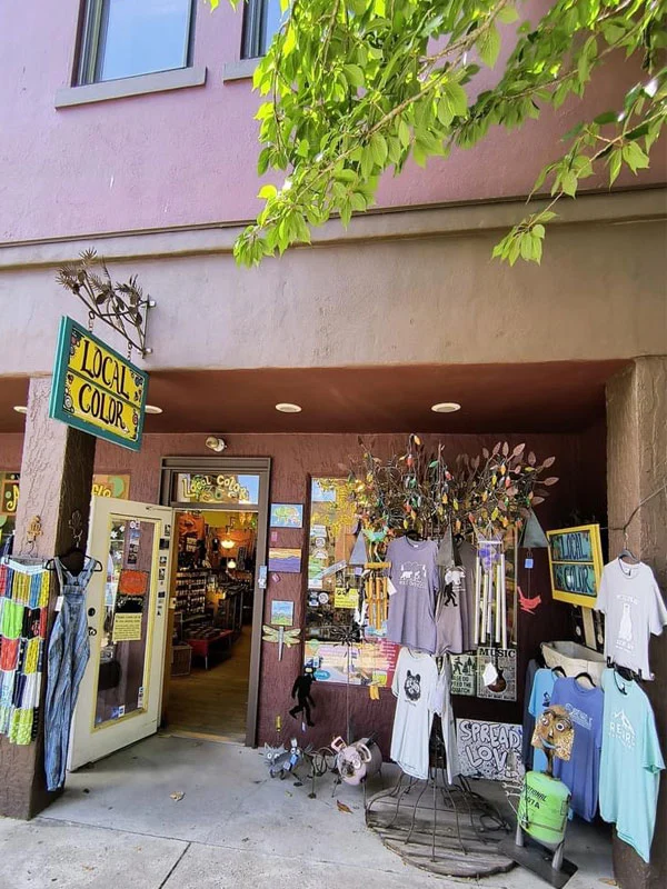 An eclectic shop named "Local Color" with a lavender exterior displays various colorful items outside, including T-shirts, art, and decorations. An open door reveals the inside of the store, which is filled with more items. Trees with green leaves frame the top of the image.