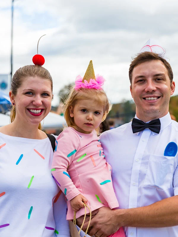 A smiling family at an outdoor event: a woman in a white shirt with colorful sprinkle patterns, a red cherry hat, a pink-clad child with a gold unicorn horn hat, and a man in a white shirt, bow tie, with a blue ribbon and ice cream hat.