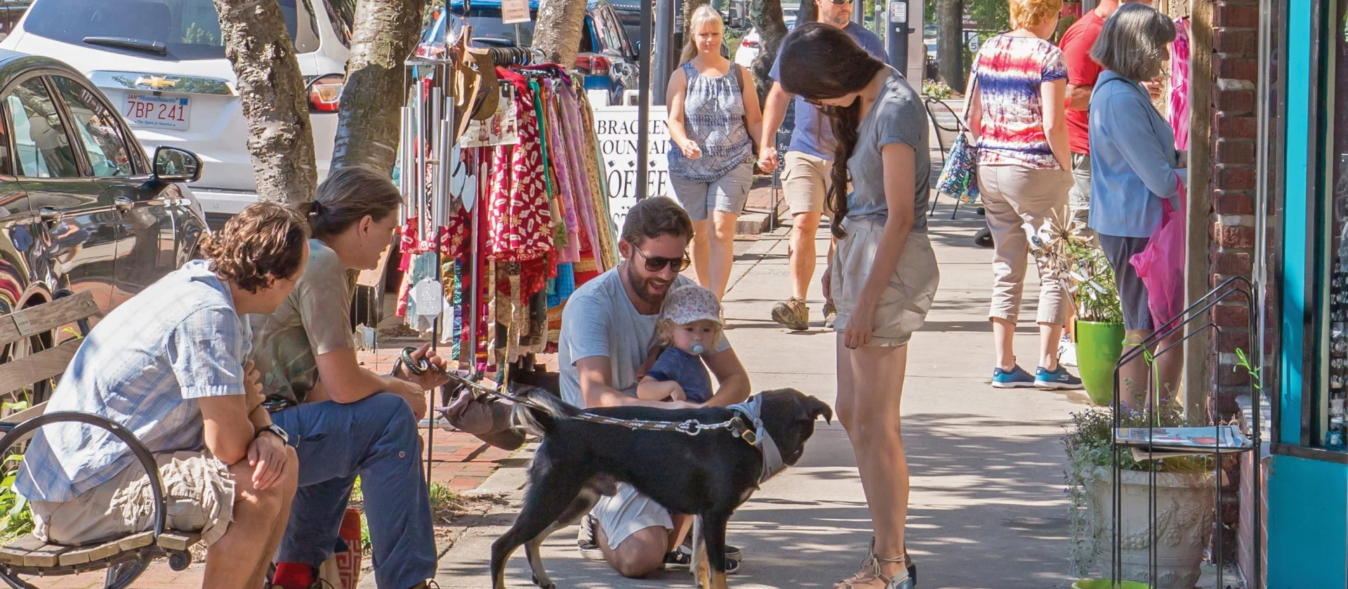 A lively street scene shows several people interacting. Two men are seated on a bench, while a person kneels to pat a black dog held by an adult standing with a child in a stroller. Several colorful clothes hang on racks nearby, and other pedestrians walk by.