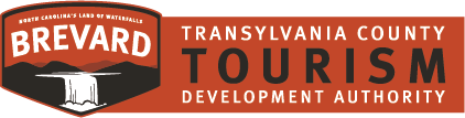 Logo of the Brevard/Transylvania County Tourism Development Authority. The left side features a shield with the text "Brevard" and "North Carolina's Land of Waterfalls" above a stylized waterfall. The right side has text reading "Transylvania County Tourism Development Authority.