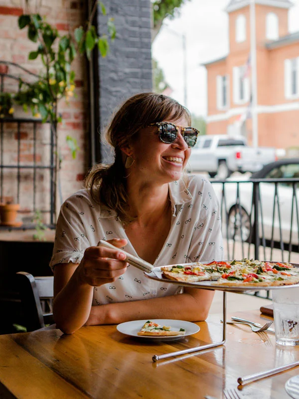 A woman sits at a wooden table indoors by a window, smiling brightly while wearing sunglasses. She is holding a pizza slicer and appears about to take a slice of pizza from a raised pizza tray in front of her. Plants and natural light fill the space.