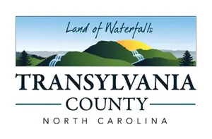 A logo for Transylvania County, North Carolina, featuring a mountainous landscape with waterfalls and a setting sun. The text reads "Land of Waterfalls" above the mountains and "Transylvania County North Carolina" below.