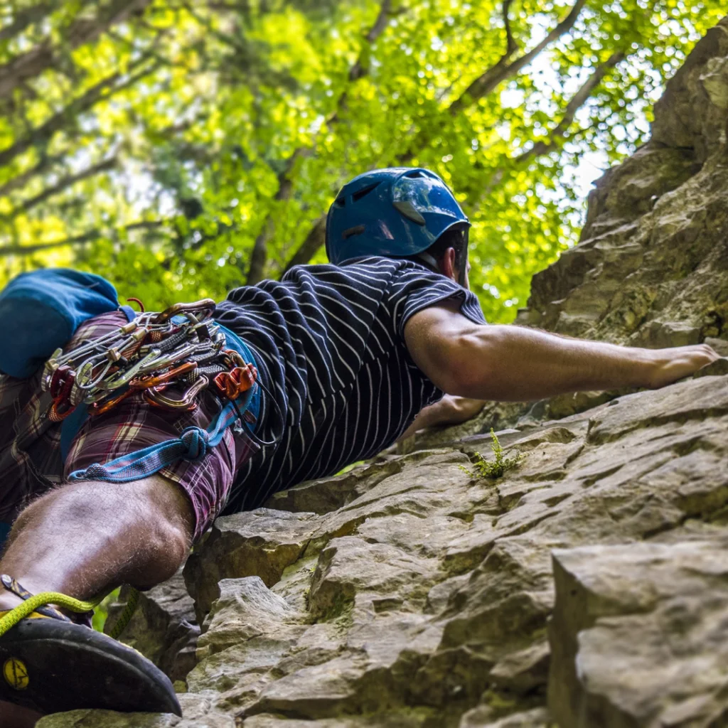Photo of a rock climber wearing a purples shirt blue helmet climbing a rock face. Photo is taken from below looking up close to the climber.