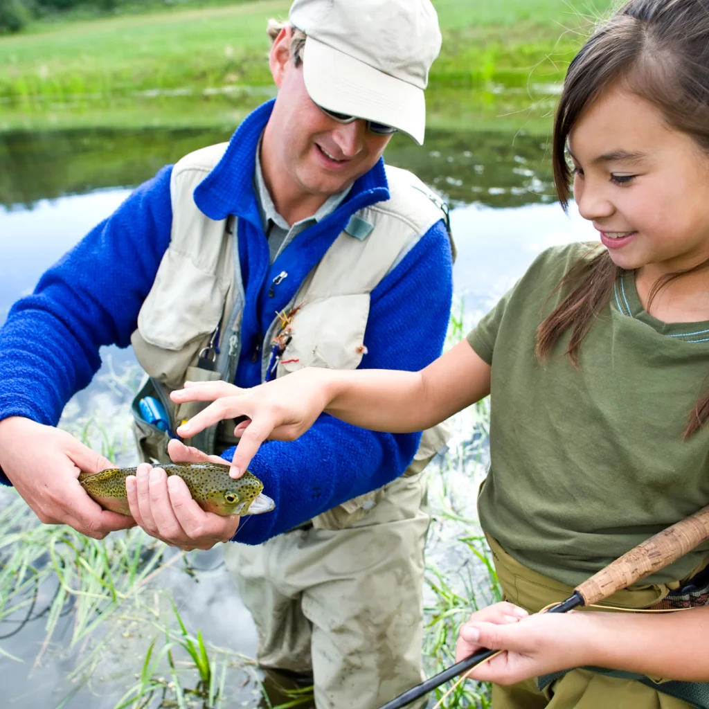 Photo a man wearing waders, a vest and a hat standing a creek hold a trout out so his daughter standing next to him can touch it.