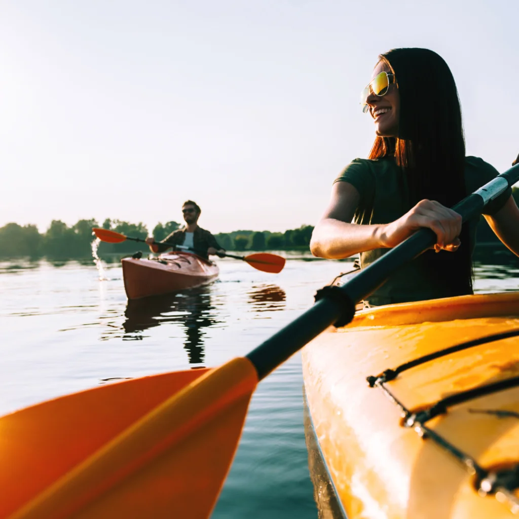 Photo of a woman in a yellow sea kayak on a lake with a male in the background behind her in another kayak. Photo is taken from the bow of her boat.