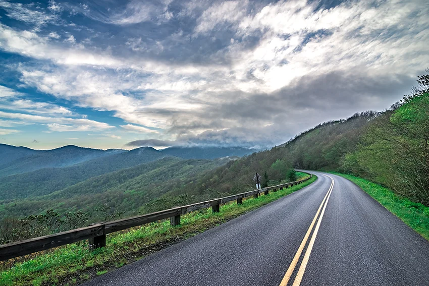 Photo of the Blue Ridge Parkway with mountain in the background and on the right and a valley to the left. There is a partly cloudy sky in the background with the clouds lit up by sunlight.