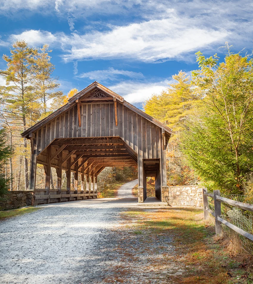 Photo of a gravel road in Dupont State Forest leading to an old covered bridge crossing a river surrounded by trees in fall covers and blue sky with some clouds.