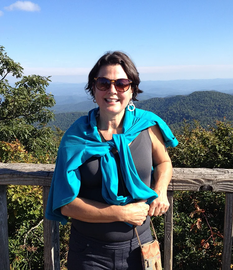 Photo of Ann Hollingsworth leaning against a wooden railing with the view of mountains in the background on the Blue Ridge Parkway.