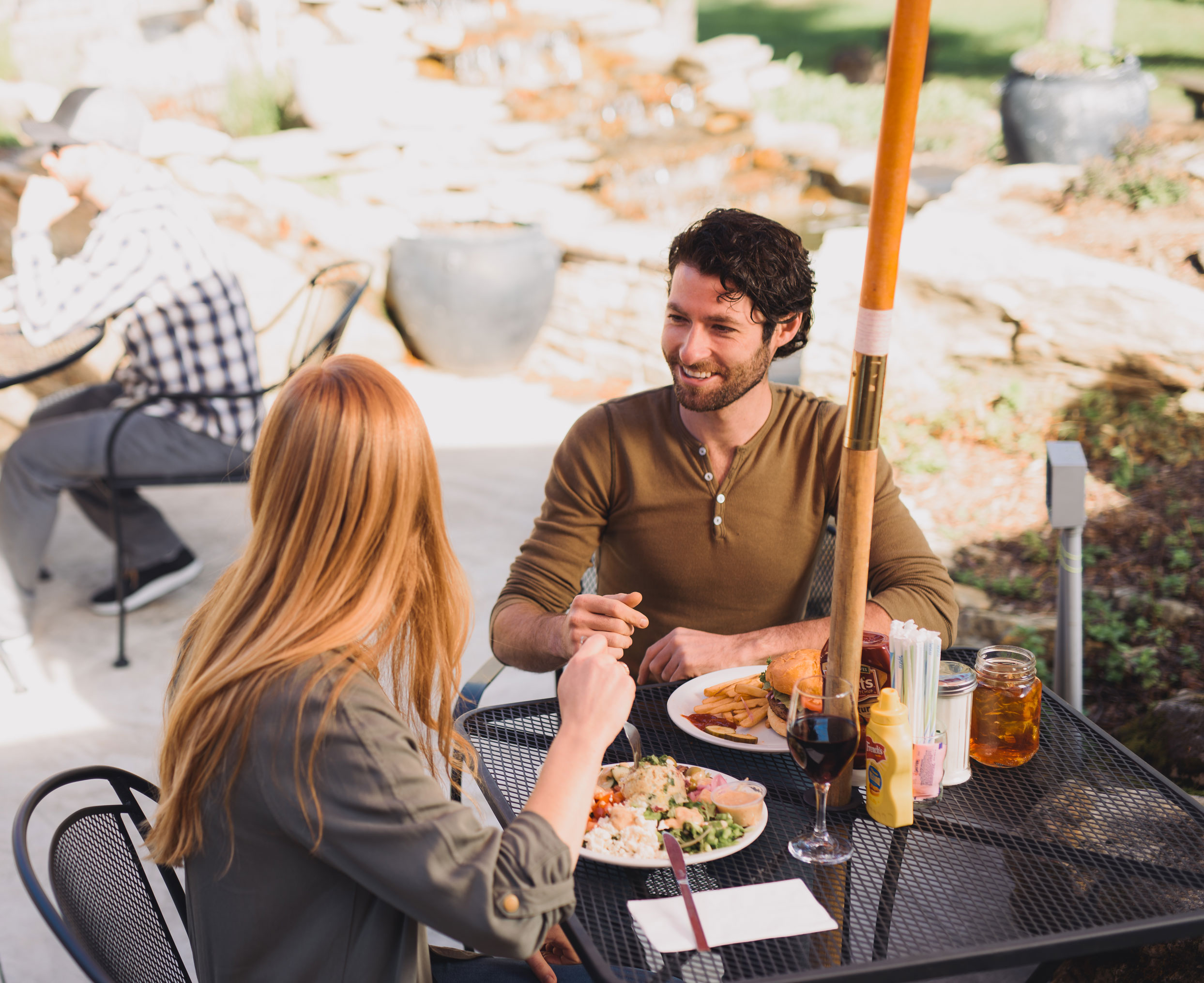 A beautiful stone house with a gray shingled roof and a chimney. The front entrance has white columns and an arched doorway surrounded by lush green bushes and trees. The house has several dormer windows and a well-maintained lawn in front.