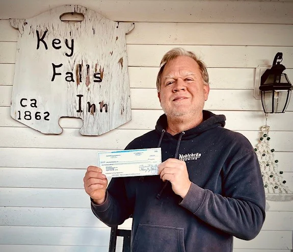 Photo of Richard Bush holding a check in front of a Key Falls Inn with a Key Falls Inn sign behind him on the wall.